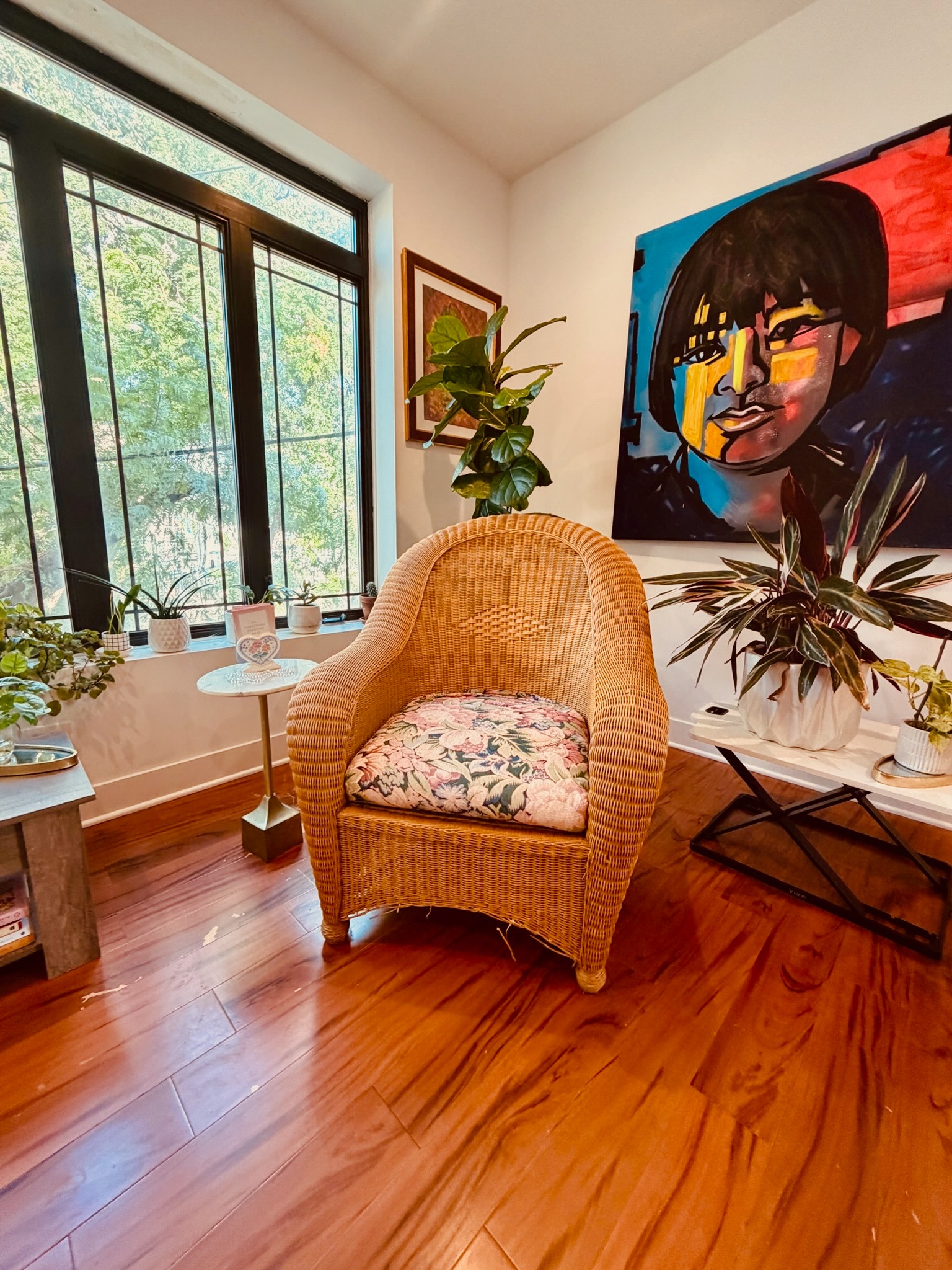 Wicker chair beside a sunlit window, surrounded by plants and colorful artwork
